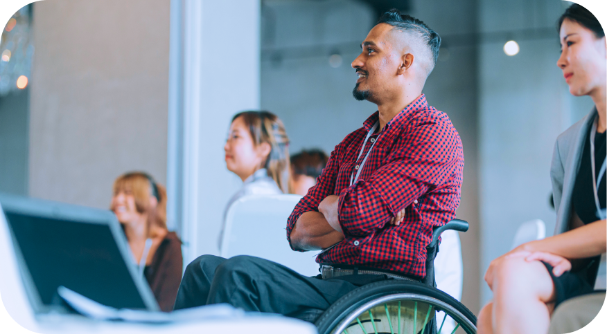 A man sitting in a wheelchair is smiling while engaging with the presentation in front of him. He is wearing a red checkered shirt and is surrounded by several attendees, some of whom are partially visible in the background, appearing to listen to the event. A laptop is positioned to the left, suggesting a work or conference setting.
