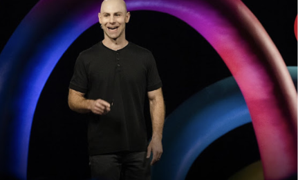 A man with a shaved head wearing a black shirt smiles while standing in front of colorful, curved abstract shapes on a dark background, suggesting an engaging presentation or speech.