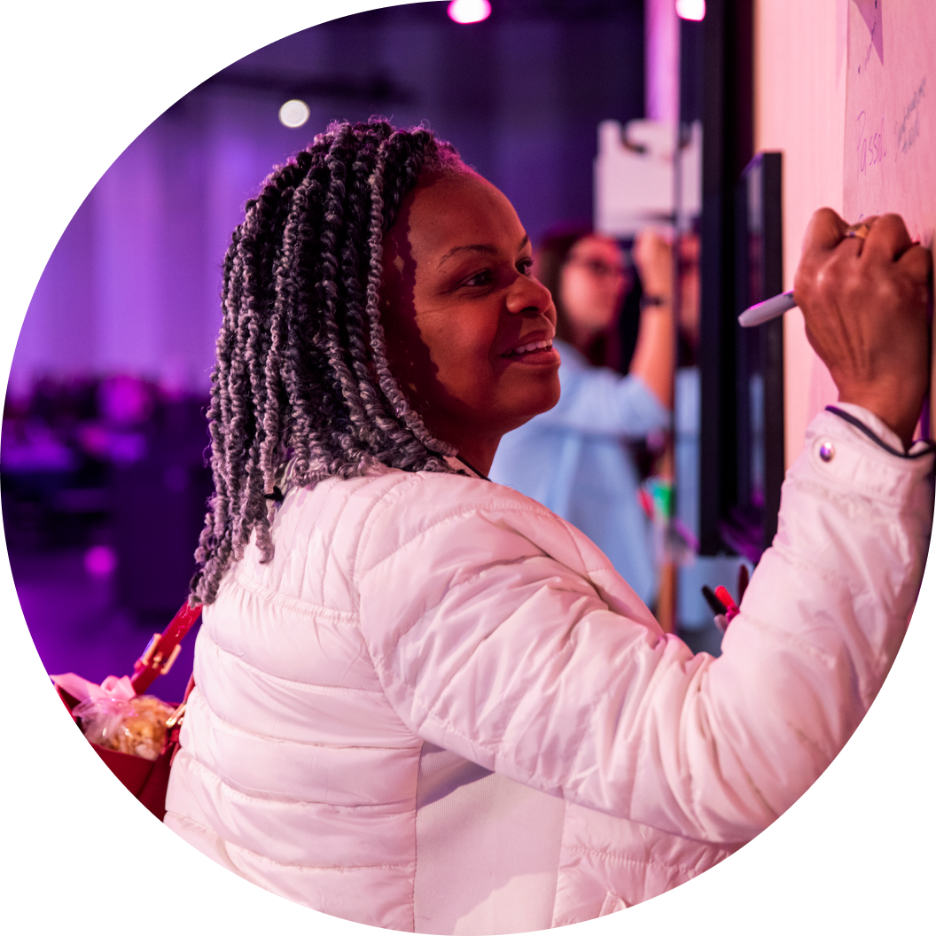 A woman with braided hair smiles as she writes on a whiteboard with a marker during an event. In the background, another person can also be seen writing. The setting features a vibrant atmosphere with purple lighting. The woman appears engaged and happy in the moment.