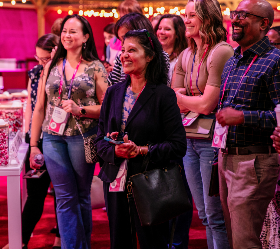 A group of attendees smiling and engaging with each other at an event, standing in front of a table filled with treats. They are dressed casually and wearing name tags. The colorful decorations and warm lighting create a lively atmosphere.
