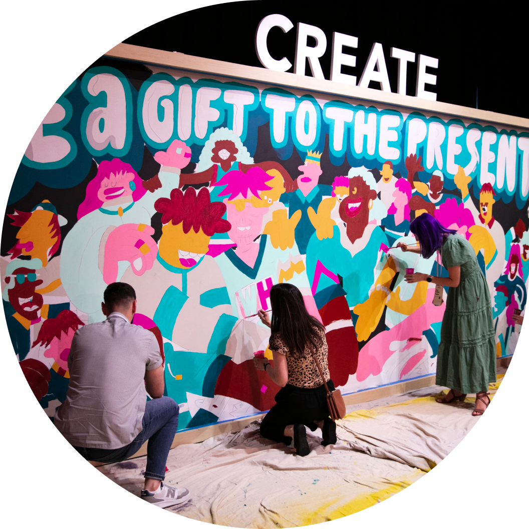 A colorful mural featuring diverse, stylized human figures is being painted by three individuals. In the foreground, two people are working on the mural; one is sitting and applying paint, while another is kneeling and focused on adding details. A large sign above the mural reads "CREATE" alongside the phrase "BE A GIFT TO THE PRESENT," suggesting a theme of creativity and expression. The overall atmosphere is vibrant and engaging, with a bright palette of colors that highlights the artistic collaboration.