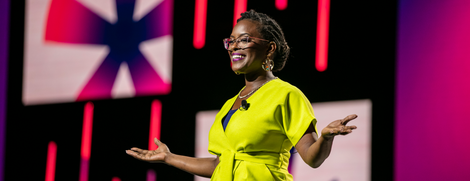 A woman smiling and gesturing with her hands is speaking on stage, wearing a bright yellow outfit. Behind her are colorful screen displays with abstract shapes, including a star-like pattern and vertical lines. The setting suggests a lively presentation or event.