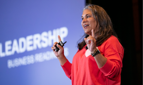 A speaker in a red sweater is gesturing with both hands while addressing an audience at a conference. Behind her, a screen displays the word 