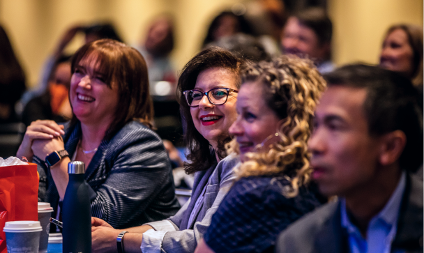 A group of smiling individuals engage attentively in a lively setting, likely during a conference or event. In the foreground, two women are prominently featured—one with glasses and the other with curly hair—while a man is visible on the right. The background shows several attendees, suggesting a larger audience. Various items such as cups and bags are placed on the table in front of them, contributing to the vibrant atmosphere.