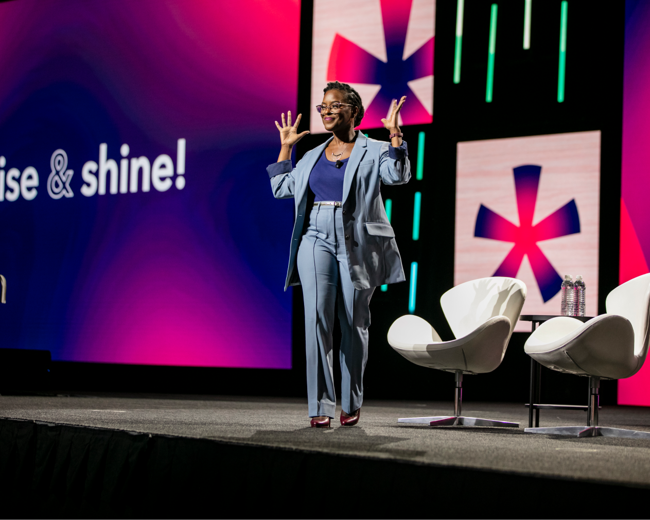 A woman enthusiastically gestures with her hands on stage, wearing a blue suit and smiling. Behind her, a large screen displays the text 