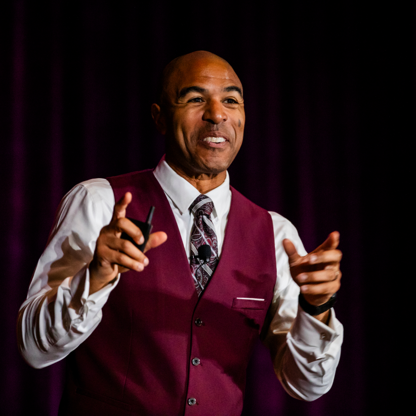 A man with a warm smile is presenting on stage, wearing a burgundy vest over a white shirt and a patterned tie. He holds a small device in one hand, gesturing animatedly as he engages with the audience. The background features a deep purple curtain, creating a professional yet inviting atmosphere for the presentation.