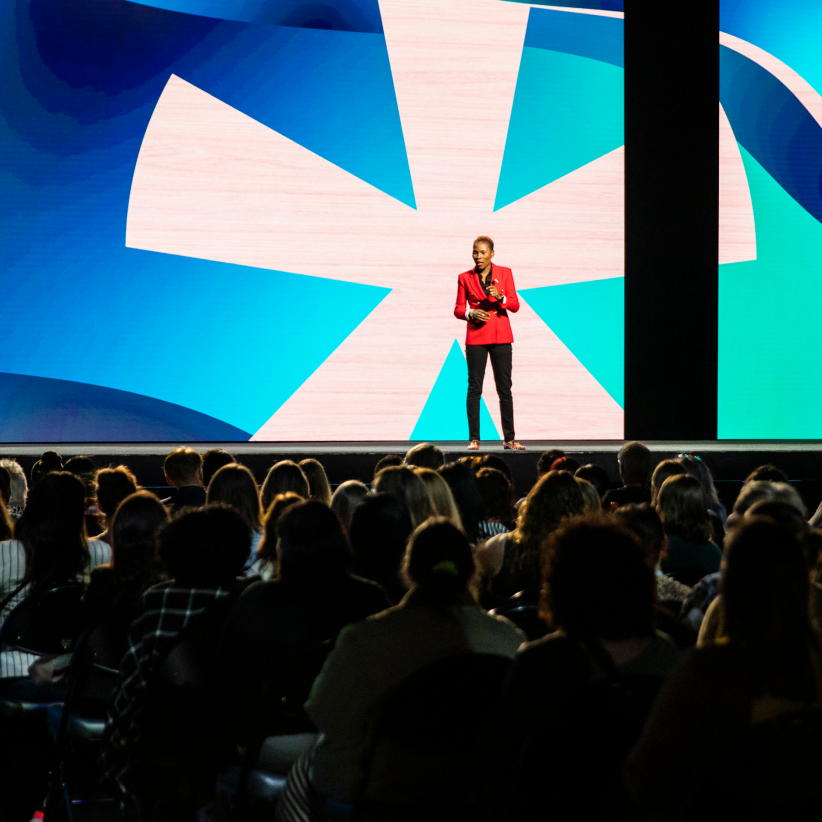 A speaker in a bright red blazer stands on stage in front of a large screen displaying abstract shapes in blue and white. The audience, made up of attendees seated in rows, is focused on the speaker, who appears to be holding a microphone.