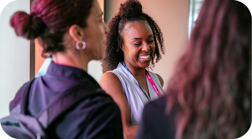 A group of two women converses, with one woman smiling while wearing a badge around her neck. The background features muted colors, enhancing the friendly atmosphere of the interaction. The women appear engaged and happy in their discussion.