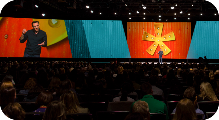 A speaker gestures animatedly on stage during a presentation, with a large screen behind him displaying a colorful visual featuring a yellow starburst design against a backdrop of wood textures. The audience, consisting of many seated individuals, attentively watches the speaker. Bright stage lights illuminate the scene, creating an engaging atmosphere.