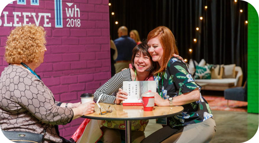 Two women smiling and embracing at a table, holding a book titled "Change the World" while sitting in a lively conference environment. One woman has curly blonde hair and wears a patterned top, while the other has straight black hair and is dressed in a floral shirt. A coffee cup and additional cups are on the table, and colorful decorations and soft furnishings are visible in the background. The wall behind them displays the text "wh 2018."