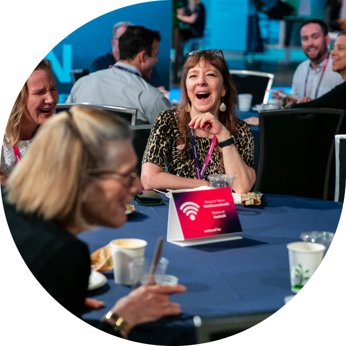 A group of attendees enjoying a meal at a conference table, with one woman laughing joyfully in the foreground. The table is set with drinks and snacks, and a sign that reads “Network Team” is placed centrally on the table, indicating it is part of a session or discussion. Other attendees can be seen in the background, engaging in conversation.