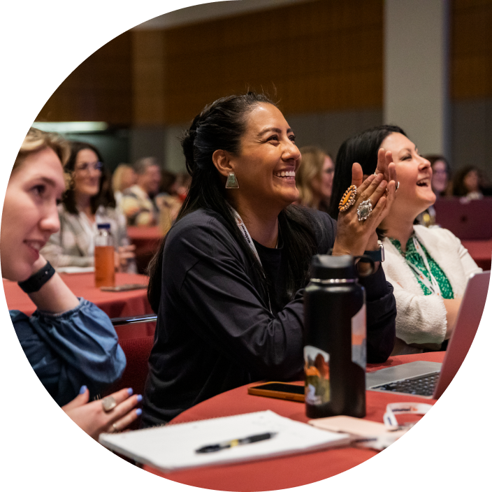 A group of attendees at a conference are engaged and smiling, with one woman prominently applauding. The setting features tables adorned with notebooks and laptops, while individuals in the background appear attentive and enjoying the moment.