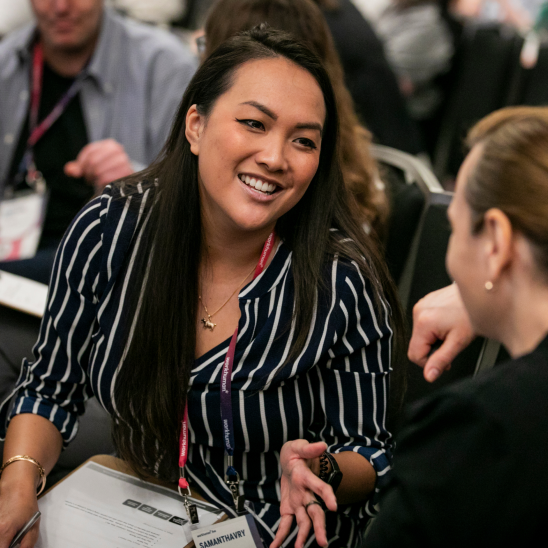 A woman with long, dark hair wearing a striped shirt smiles while engaged in a conversation with another person. They are seated at a table in a busy event setting, with attendees visible in the background. The woman has a name tag visible, suggesting an organized gathering or conference atmosphere.